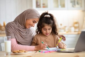 Loving Muslim Mom In Hijab Drawing With Her Little Preschooler Daughter In Kitchen, Happy Islamic Family Sitting At Table And Using Colorful Pencils, Having Fun Together At Home, Free Space