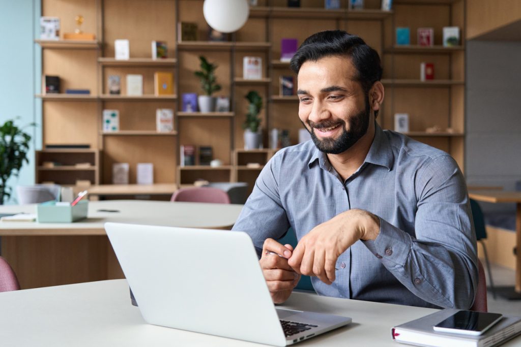 Happy smiling Indian businessman having online virtual meeting call on laptop looking at screen sitting at table in coworking creative office, professional manager talking to employees.