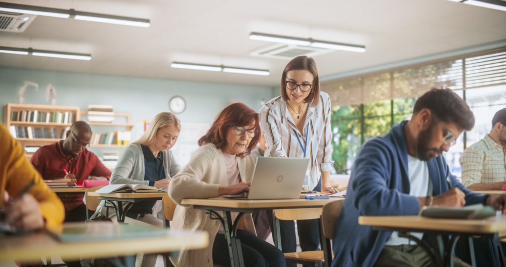 Young Female Teacher Giving a Lecture During an Adult Education Course in School, Having a Conversation with a Older Female with Laptop. Diverse Mature Students Doing Textbook Exercises in Classroom