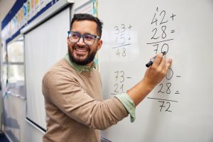 Happy teacher teaching how to do the column additions at elementary school. Smiling man explaining additions in column in primary class while writing on whiteboard. Maths teacher explaining arithmetic sums to elementary children.