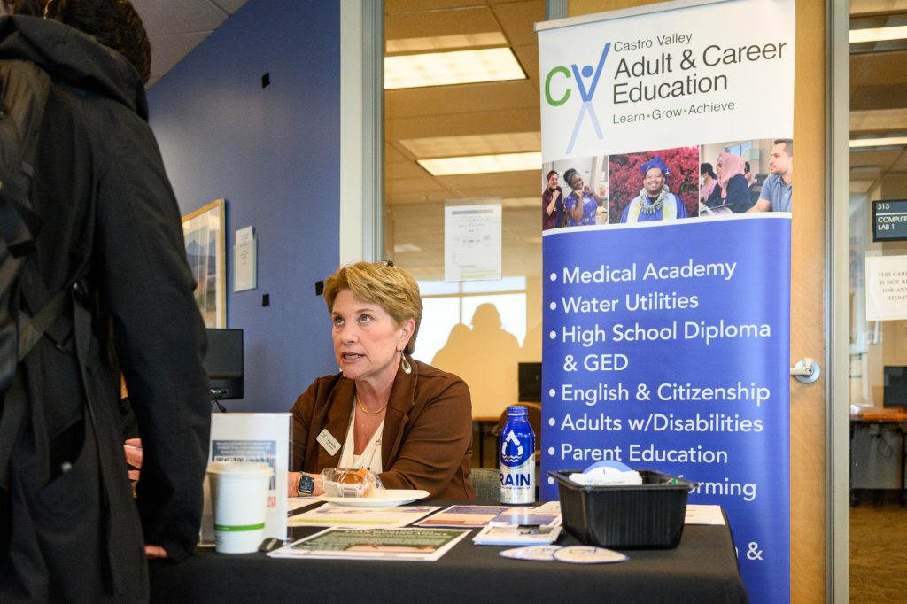 Jamie Renton sitting at a booth for Castro Valley Adult Education, speaking with a man