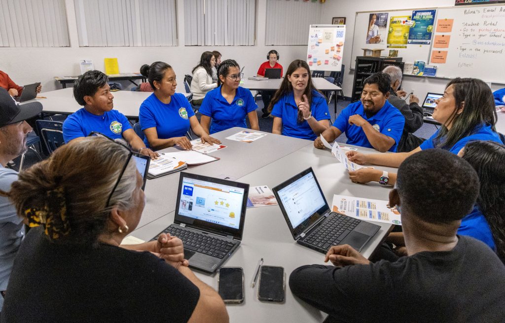 A group of adult students sitting around a table, talking. All dressed in a blue polo uniform.