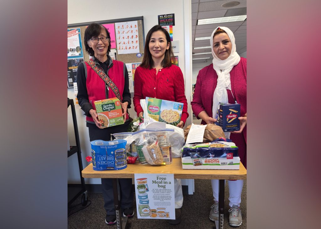 Three women posed, standing in front of a table containing food for a food drive.
